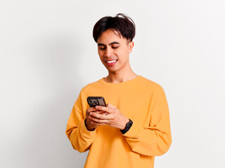 Fototapeta premium A cheerful young Asian man wearing a yellow sweatshirt smiles while holding and typing on his smartphone.