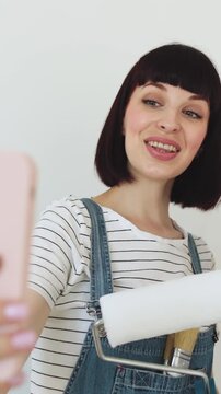 A mother and her young daughter are taking a selfie while sitting on a ladder with paint rollers, preparing to redecorate a room