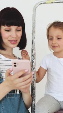 A mother and her young daughter are on a ladder, looking at a phone while preparing to paint a room