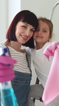 A smiling mother and her daughter are cleaning a room together, holding cleaning supplies and a cloth