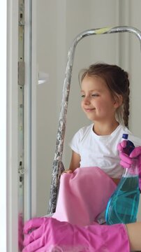 A mother and her young daughter wearing pink gloves clean a window together with a spray bottle and cloth