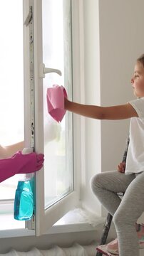 A mother and her young daughter work together to clean a large window, with the child standing on a ladder to reach higher spots