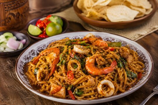 Traditional Acehnese Mie Aceh Seafood Pedas with Pickles, Emping and Lime on Rustic Wooden Table