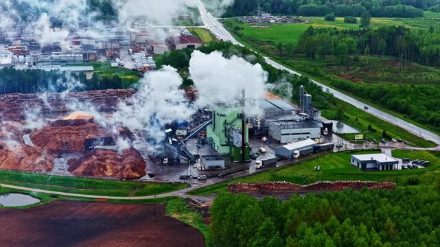 Aerial view of Stora Enso sawmill with steam rising from chimney beside huge piles of wood chips and sawdust surrounded by green forest in rural Latvia