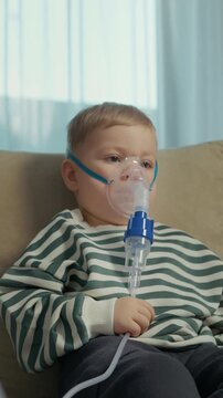 Vertical Screen: A child is sitting on a couch using a nebulizer with a mask to inhale a saline solution for treating cough and other respiratory problems at home..