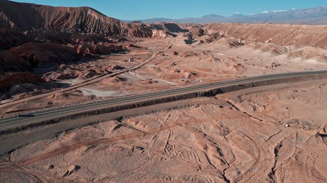 Backward flight over paved highway cutting through dramatic eroded salt rock badlands revealing cracked desert floor and snow-capped Andean peaks beyond. Valle de la Muerte, Chile