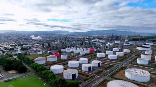 Aerial view over vast oil refinery complex with rows of white and red storage tanks distillation towers and smoke stacks with residential city and mountains in the background. Hualp&eacute;n, Biob&iacute;o, Chile