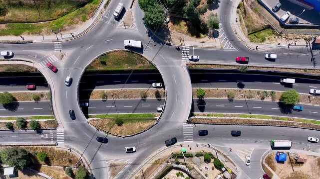 Top down dolly aerial shot over urban roundabout overpass with cars flowing through circular ring road above busy highway underpass with crosswalks and surrounding streets
