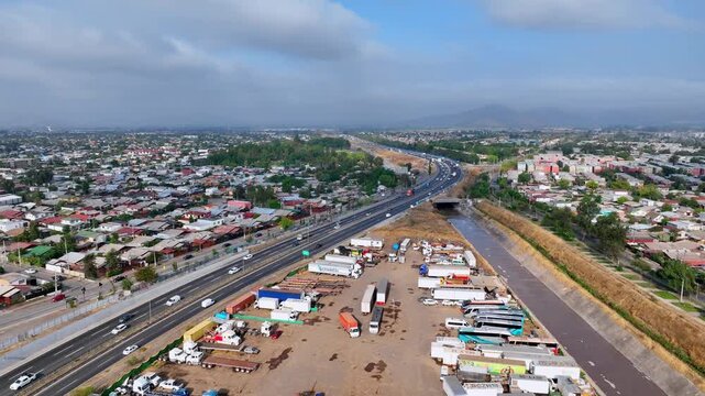 Forward flight over Maip&uacute; highway corridor with busy truck depot dry river channel and dense residential neighborhoods stretching toward Andean foothills under dramatic cloudy sky. Maip&uacute;, Santiago, Ch