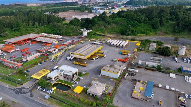 Tilt down aerial shot over liquefied natural gas distribution plant with orange and yellow facilities truck fleet loading bays and port infrastructure visible along the Pacific coastline. Hualpen, Bio