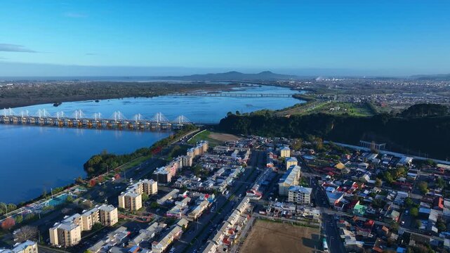 Dolly shot over Concepcion riverside neighborhoods revealing the iconic cable stayed Bio Bio bridge spanning the wide calm river with distant hills under vivid blue sky. Concepcion, Chile