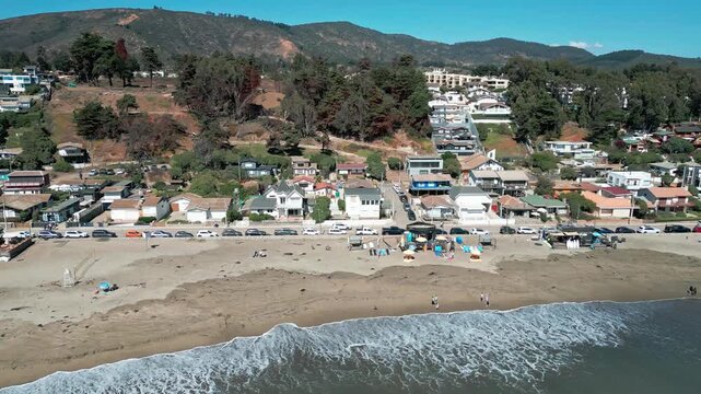 Sliding reveal over Maitencillo beachfront showing sandy shore people on the beach coastal village buildings parked cars and forested hillside under clear blue sky. Maitencillo, Valparaiso, Chile