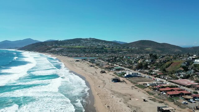 Gliding flight along Maitencillo coastline with powerful Pacific surf breaking on wide sandy beach beside coastal village and dry rolling hills under vivid blue sky. Maitencillo, Valparaiso, Chile