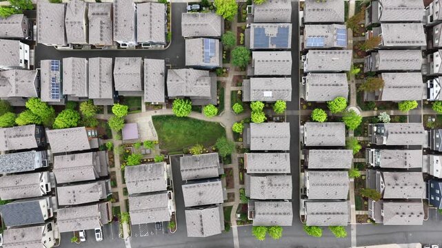 Beaverton residential complex from directly above with uniform rooftops, solar panels, grid pathways, green courtyard, trees and street parking under overcast sky. Oregon, USA. Aerial top down shot.