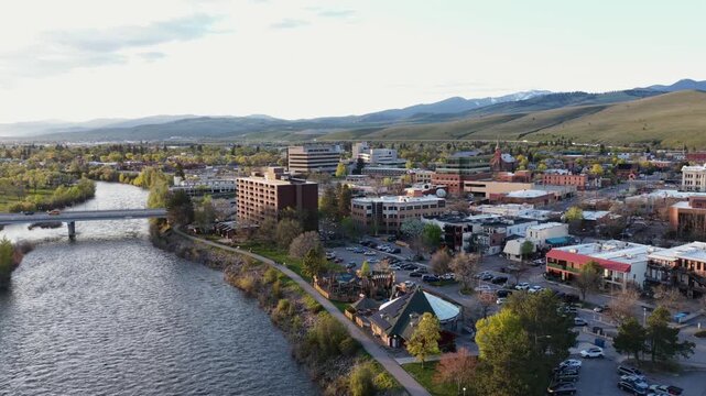 Missoula downtown waterfront with Clark Fork River, riverside walking path, commercial and brick buildings, parking lots and snow-capped rolling hills under evening sky. Montana, USA. Aerial wide shot