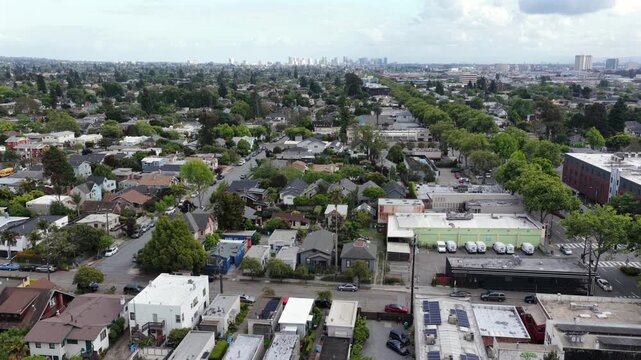 Dense residential neighborhood with tree-lined streets, mixed housing and commercial buildings stretching toward distant downtown skyline under partly cloudy sky. California, USA. Aerial descend shot.