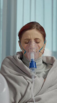Vertical Screen: A woman is using a nebulizer with a mask to inhale a saline solution for her cough. She is sitting indoors while treating her respiratory infection.
