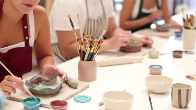Girl paints bowl of clay during master class, top view. Unrecognizable people team of creative enthusiasts is engaged in clay modeling lesson. Employee covers gray, damp clay bowl with paint