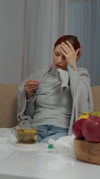 Vertical Screen: A woman sits on a sofa feeling unwell. She checks her temperature with a thermometer. Tea, fruit, and napkins are nearby as she tries to recover from her cold.