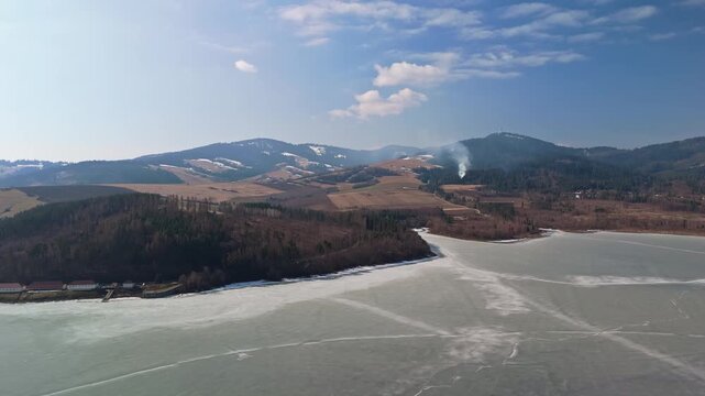 In northern Slovakia, the frozen Orava Reservoir extends beside snow‑patched fields, coniferous forest, and surrounding hills, seen across the winter landscape from a drone.