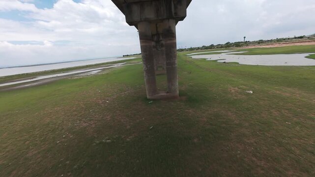 Thrilling low-altitude FPV drone shot flying under the concrete pillars of the Pa Sak Jolasid Dam railway bridge in Lopburi, Thailand. Highlights fast-paced maneuverability over green plains.