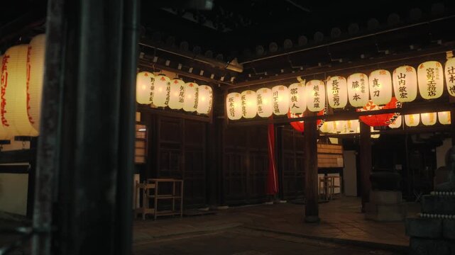 Camera pans past a wooden pillar to reveal rows of glowing chochin paper lanterns inside a Kyoto shrine at night.