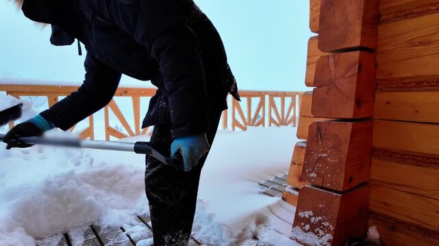 Guy shoveling heavy snow on a rustic wooden deck near a log cabin. Light snowfall adds atmosphere to this cold outdoor winter task.