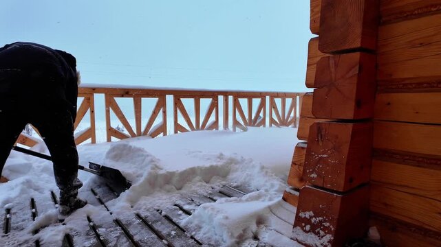 Worker in black winter gear removes thick snow from a terrace with a wide shovel.