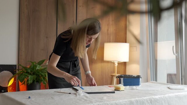 Woman creates textured canvas art at a plastic-covered table, using palette knife beside art tools. Concept of DIY wall decor and creative hobby