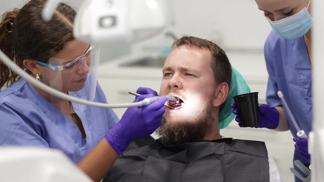 Woman dentist and a nurse carefully drill the tooth of a worried man, they remove caries and prepare the tooth for a filling. Man came to treat his teeth