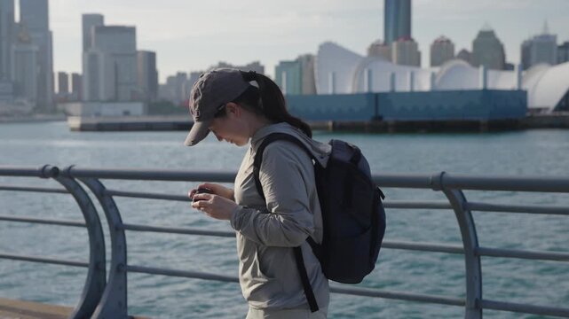 A smiling young adult female traveler wearing casual outdoor clothing and a backpack uses a compact camera to take photos at a coastal waterfront with a city skyline in the background.