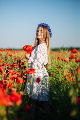 Obraz premium Blonde slavic female posing in white dress, blue flower crown, holding red poppy bouquet amid sunlit summer meadow