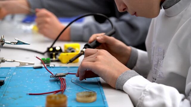 Young hands hold soldering iron with focus. Blue mat holds wires, resistors, and tools. Sweat beads show concentration and effort. Scene captures real-time electronics repair. Perfect for education