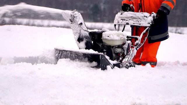 Snowblower operator clears deep snow with focused determination. Bright orange gear contrasts sharply with white surroundings. Machine chews through snow with powerful mechanical force