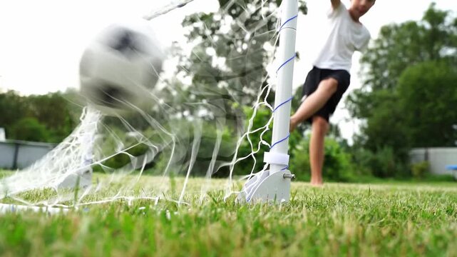 Child kicks ball toward goal on grassy field. Goal net stands ready for the next shot. Sunlight glows over the green playing area. Joyful motion suggests a fun, active moment