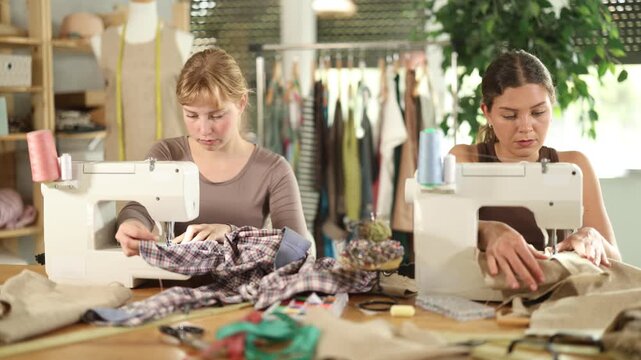 Two female tailors are sitting at a table in the workshop and sewing clothes on machines. Employees of the sewing industry make wear according to individual measurements. High quality 4k footage