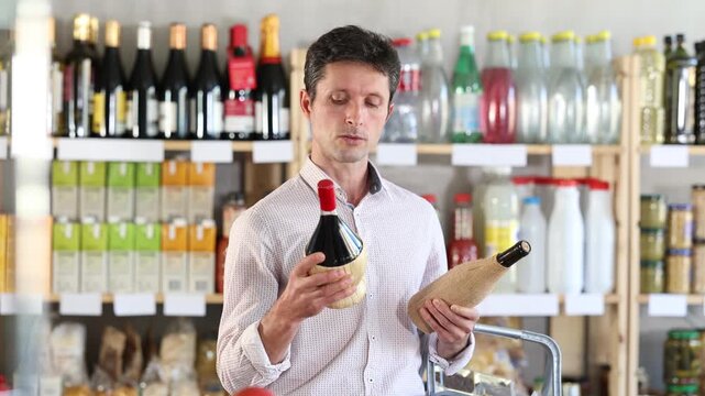 Man in light shirt took some varieties of wine from shelf. Client compare heady and new red wine items package