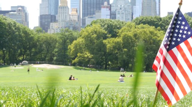 American flag on green grass in Central Park, New York, USA. Sheep Meadow, high-rise buildings and people. Skyline cityscape, Manhattan Midtown skyscrapers. United States symbol and business district