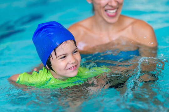 Little boy is held and guided by a female instructor while kicking through water in an indoor pool, practicing body position and leg propulsion during an early aquatic lesson.