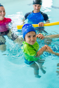 Little boy in a blue swim cap practices water movement assisted by two instructors in an indoor pool, building aquatic independence and physical coordination through early structured swimming lessons.