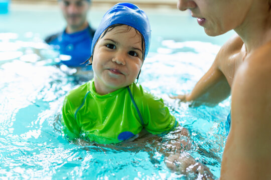Toddler boy in a blue swim cap and green vest stands in an indoor pool, engaged in focused interaction during an early aquatic lesson.