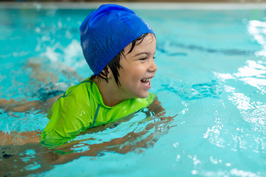 Happy boy in a blue swim cap and green flotation vest moves through turquoise pool water with visible excitement, demonstrating growing aquatic independence and comfort during swim training.