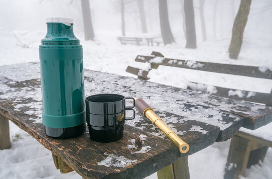 A green thermos and a black mug sit on a snowy wooden table. The scene is set in a winter landscape with fog and snow-covered benches in the background.