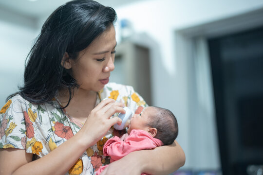 Loving Asian mother bottle-feeding her newborn baby while cradling the infant in her arms at home.