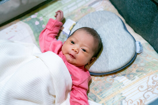 Awake newborn baby girl lying on a head-shaping pillow under a white blanket wearing pink clothes.