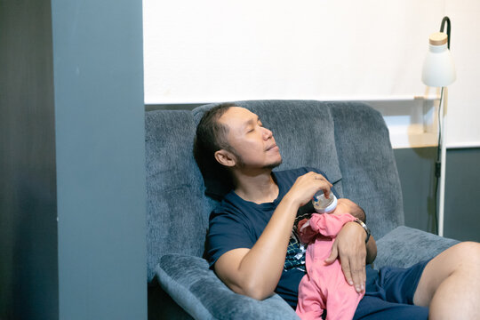 Tired father sitting on a sofa and bottle-feeding his newborn baby with his eyes closed.