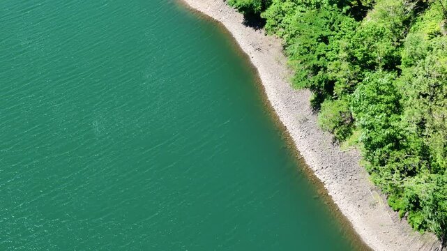 Aerial view captures calm, deep green lake waters. Rugged shoreline hugs dense, emerald forest on one side. Sunlit pebbles and stones line the water's edge gently. No people are present