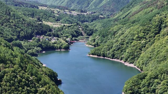 Wide river cuts through lush green hills. Vast trees surround the calm, blue waters. Meadows and distant hillsides rise gently. No people disturb the peaceful scene. Ideal for tranquil nature, travel