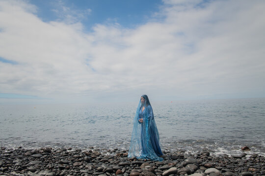 art portrait of young woman on the beach in blue dress covered by thin veil