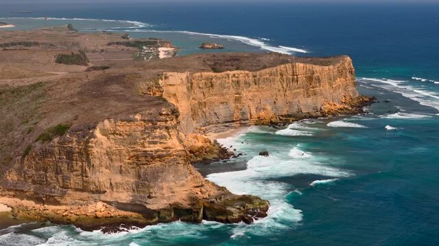 Drone ekas view captures lombok golden coastal cliffs and powerful white waves breaking along rugged ocean shoreline for travel nature geology and dramatic seascape projects